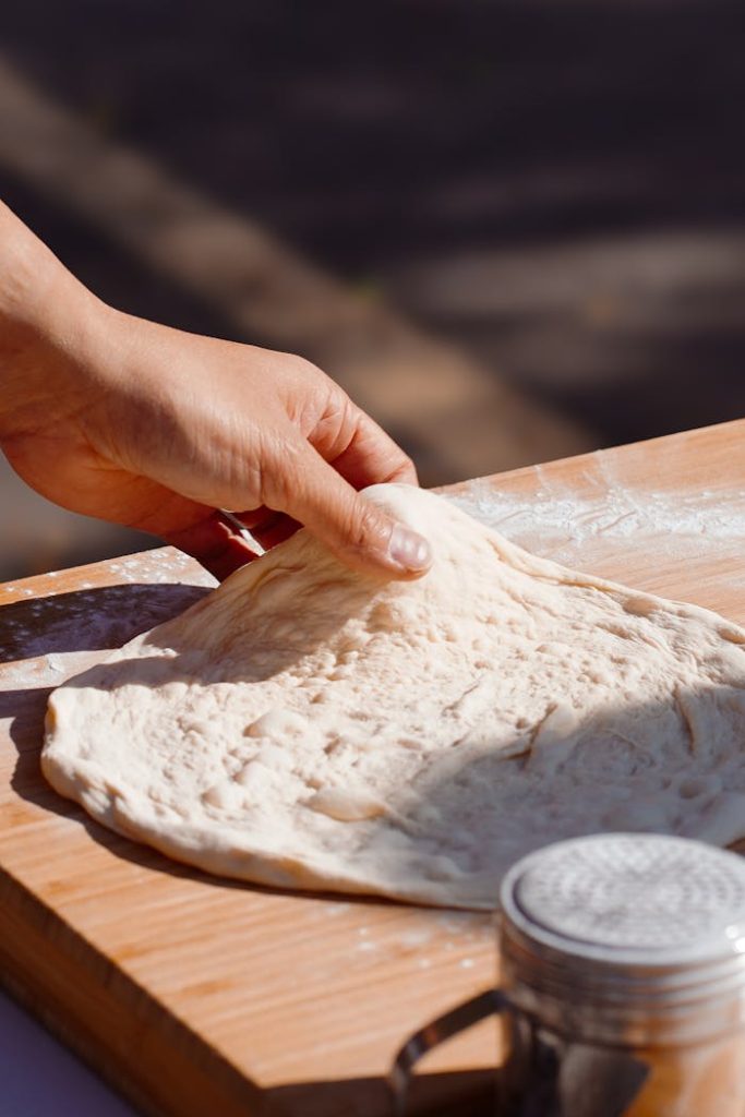 pexels-photo-28866007 Close-up of a hand shaping artisan pizza dough on a wooden board in natural light.
